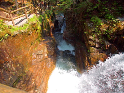 [A huge wall to the left has a wooden railing along the edge and a pathway beside it. The water comes in from the right and makes a 90 degree turn to flow away from the camera and down two or three more drops. There are lots of trees and the shade makes it difficult to tell from this distance the number of drops. The rock wall is quite wet.]